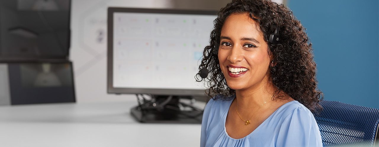 A telephone operator with headphones looks smiling into the camera, behind her are screens