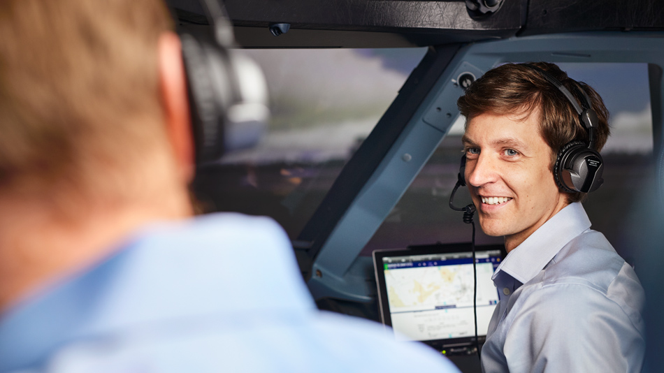 A man wearing headphones sits in an aircraft cockpit and smilingly looks at his colleague