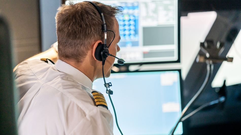 A pilot as instructor with headphones on sits in front of screens in a cockpit simulator and explains something