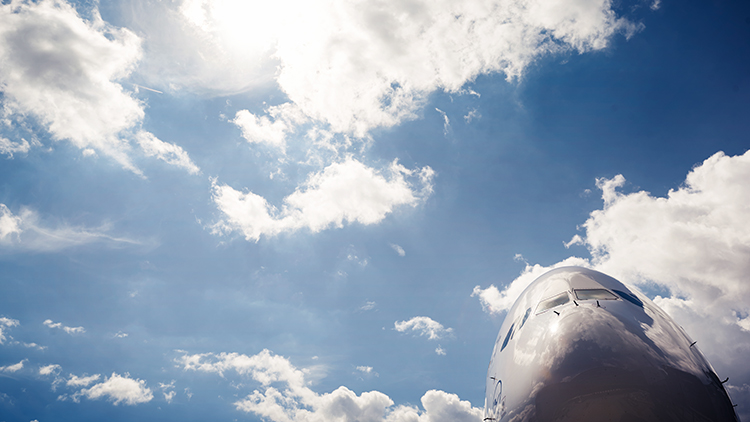 The nose of an airplane with Lufthansa logo from below in front of a blue sky with some clouds