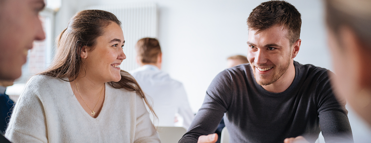 Drei Personen sitzen an einem Tisch mit einem Laptop in Business Kleidung und führen ein Gespräch.