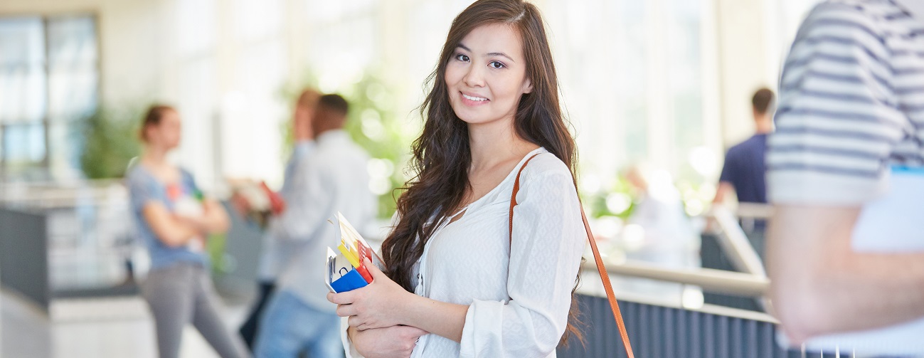 A student with a shoulder bag is holding a folder and documents in their hand and smiling at the camera.