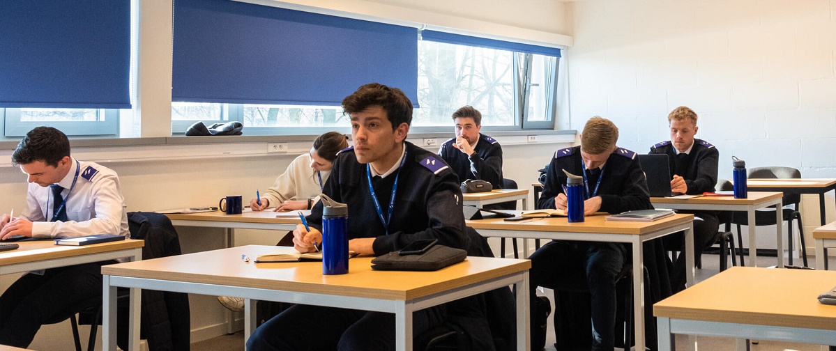 Several student pilots in uniform are sitting in a classroom, taking notes.
