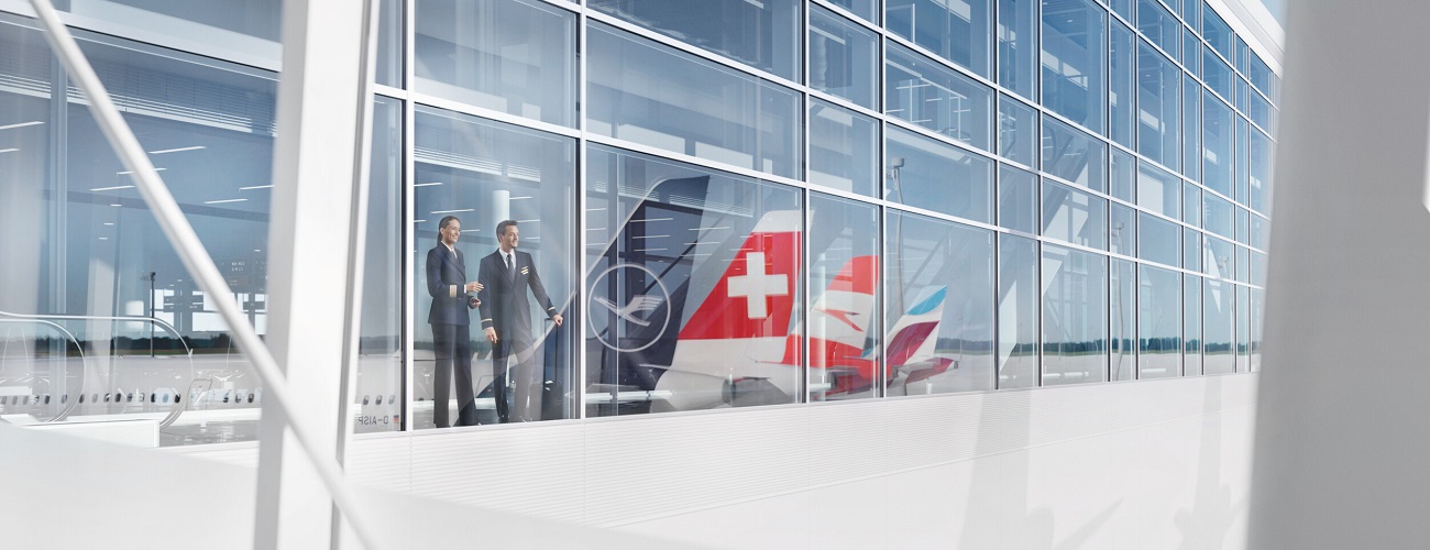 Zwei Mitglieder der Cockpit Crew stehen hinter einer verglasten Front. In den Fenster spiegeln sich die Flugzeugtails mit den Liveries von Lufthansa, Swiss, Austrian und Eurowings.