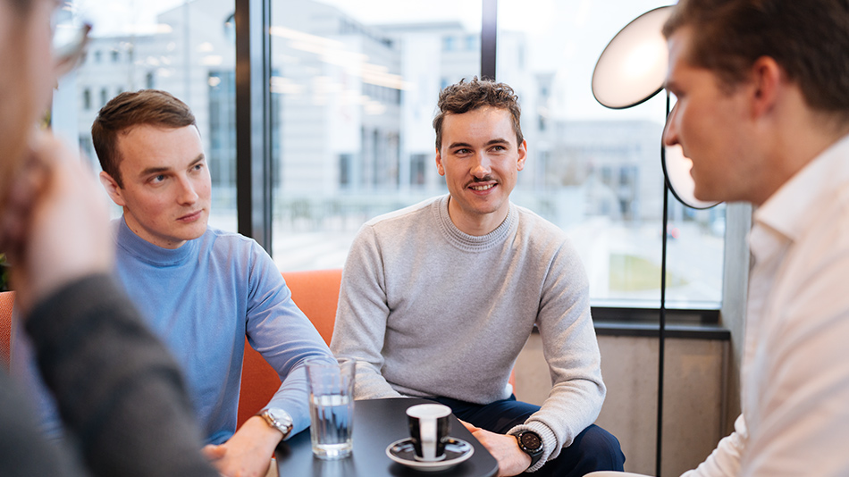 Drei lächelnde Personen stehen um einen Tisch und schauen auf einen Laptop.