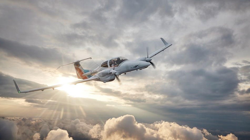 A DA42, with two people visible inside, is flying over fields and meadows under a cloudy sky.