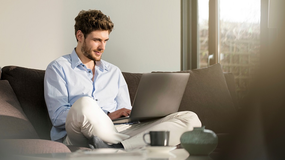 A person is sitting cross-legged on a couch with a laptop on their lap and typing on the keyboard.