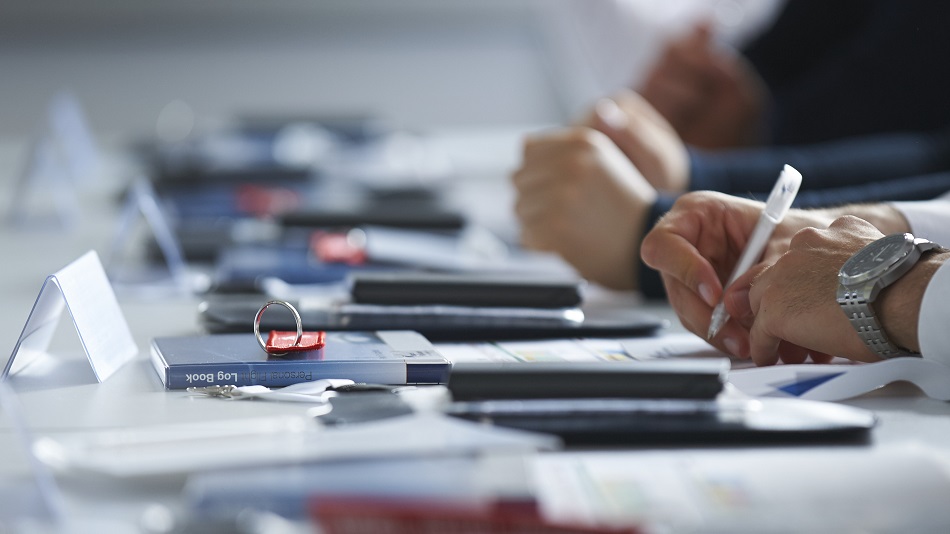 Mehrere Personen sitzen an einem Tisch und schreiben. Auf dem Tisch liegen Keyrings, Taschenrechner und Notizbücher.