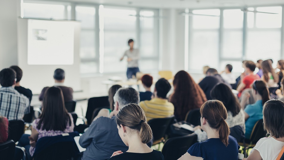 Many people sit on chairs in several rows of chairs, one person stands in front and presents.